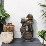 A decorative outdoor fountain with three rustic pots and a blue bird figurine features subtle LED lights, placed on a patio near a stump table, woven bag, and lush green plants.