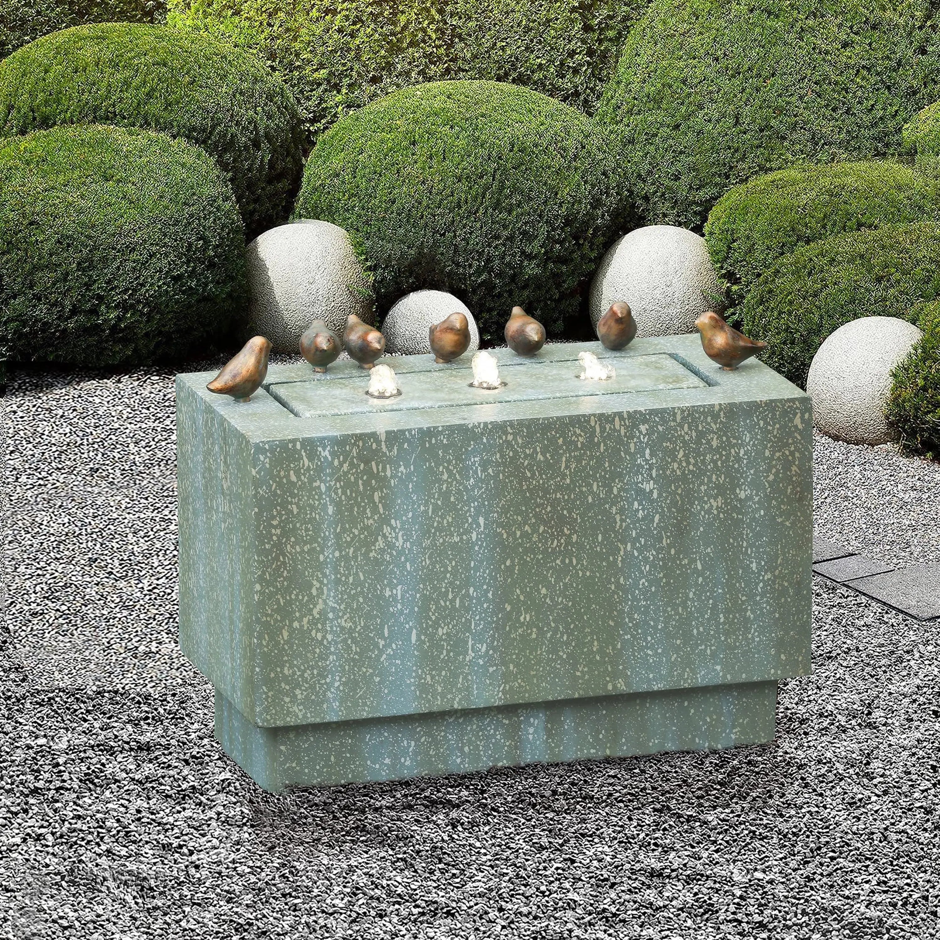 A rectangular stone birdbath with six small bird sculptures on its edges serves as a charming outdoor fountain, set in a gravel garden with trimmed green bushes and large round stones in the background.