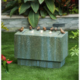Stone outdoor fountain with flowing water spouts and six small bird sculptures on top, set on grass in a garden with leafy plants in the background.