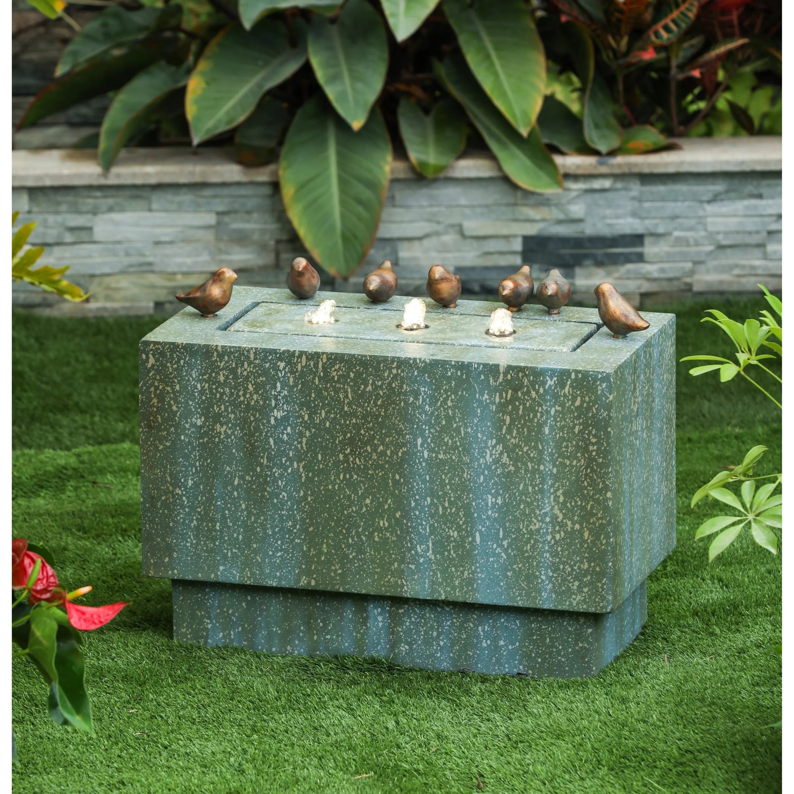 Stone outdoor fountain with flowing water spouts and six small bird sculptures on top, set on grass in a garden with leafy plants in the background.