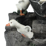A white duck figurine with an orange beak and feet sits on gray textured rocks near a small waterfall feature, adding charm to any outdoor space.