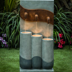 A modern fountain with LED light features three vertical cylinders and water spouts, creating a stunning focal point for outdoor living areas, set on green grass with plants and flowers in the background.