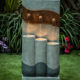 A modern fountain with LED light features three vertical cylinders and water spouts, creating a stunning focal point for outdoor living areas, set on green grass with plants and flowers in the background.