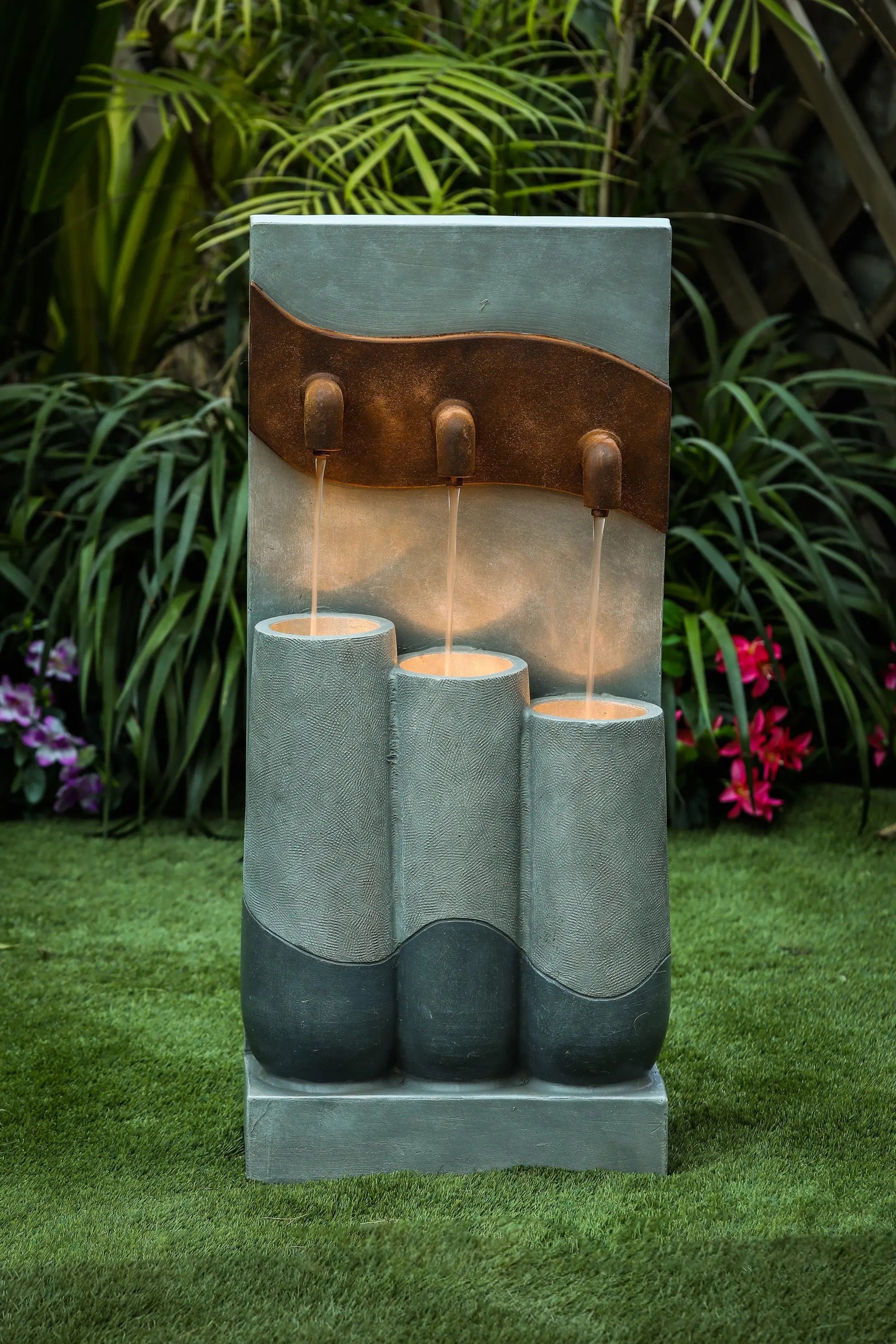 A modern fountain with LED light features three vertical cylinders and water spouts, creating a stunning focal point for outdoor living areas, set on green grass with plants and flowers in the background.
