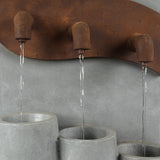 Three metal spouts with a rusted finish pour streams of water into three textured gray cylindrical basins below, creating stacked urns perfect for outdoor living areas, all set against a plain gray background.
