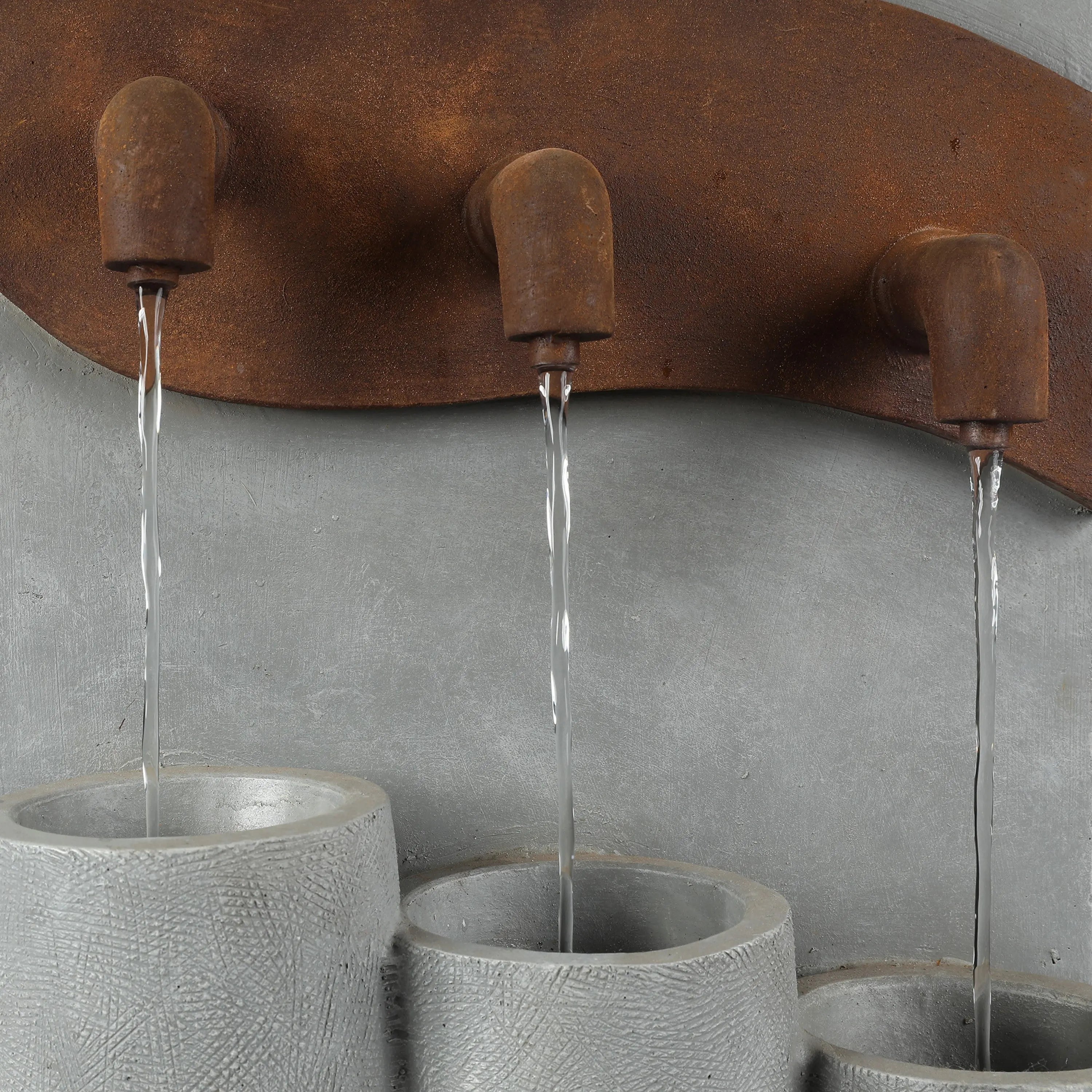 Three metal spouts with a rusted finish pour streams of water into three textured gray cylindrical basins below, creating stacked urns perfect for outdoor living areas, all set against a plain gray background.