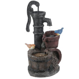A decorative farmhouse outdoor fountain with a black hand pump, two artificial birds, and water flowing into a wooden bucket and small jug adds rustic charm to any space.