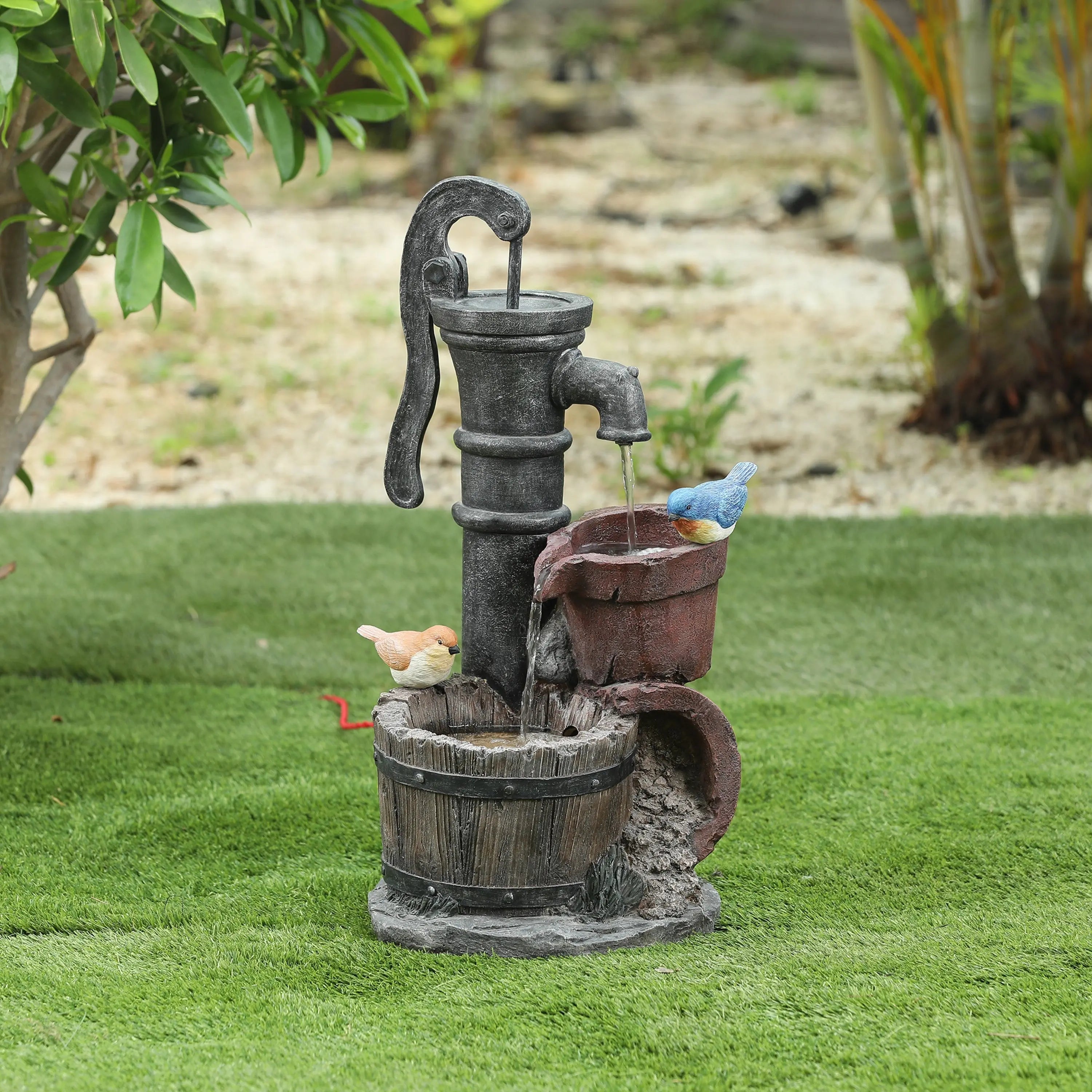 A decorative farmhouse outdoor fountain featuring a water pump with two small bird figurines, one blue and one yellow, perched on the pump and bucket, adds rustic charm to green artificial grass.