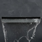 A close-up of water flowing from a modern gray rectangular spout against a dark, textured wall, capturing the elegance of a waterfall outdoor fountain.