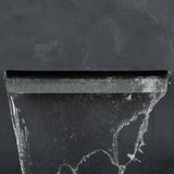 A close-up of water flowing from a modern gray rectangular spout against a dark, textured wall, capturing the elegance of a waterfall outdoor fountain.