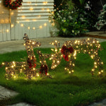 Three wire-frame reindeer, part of festive Deer Family Decorations, are adorned with lights and red bows on a grassy lawn, with more LED Holiday Decor and greenery twinkling in the background.
