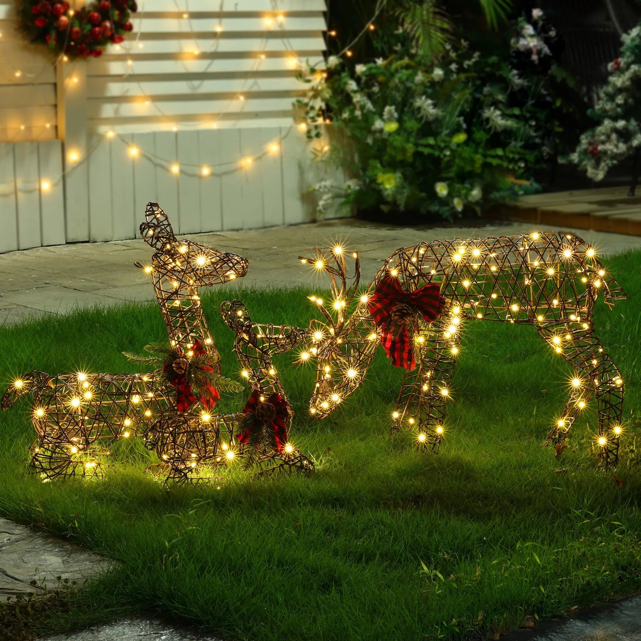 Three wire-frame reindeer, part of festive Deer Family Decorations, are adorned with lights and red bows on a grassy lawn, with more LED Holiday Decor and greenery twinkling in the background.