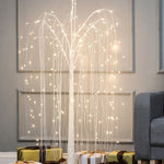 A romantic willow tree with LED lights stands next to a gray sofa, with gold and white wrapped presents arranged underneath on a wooden floor.