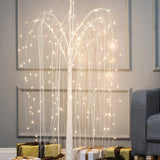 A romantic willow tree with LED lights stands next to a gray sofa, with gold and white wrapped presents arranged underneath on a wooden floor.