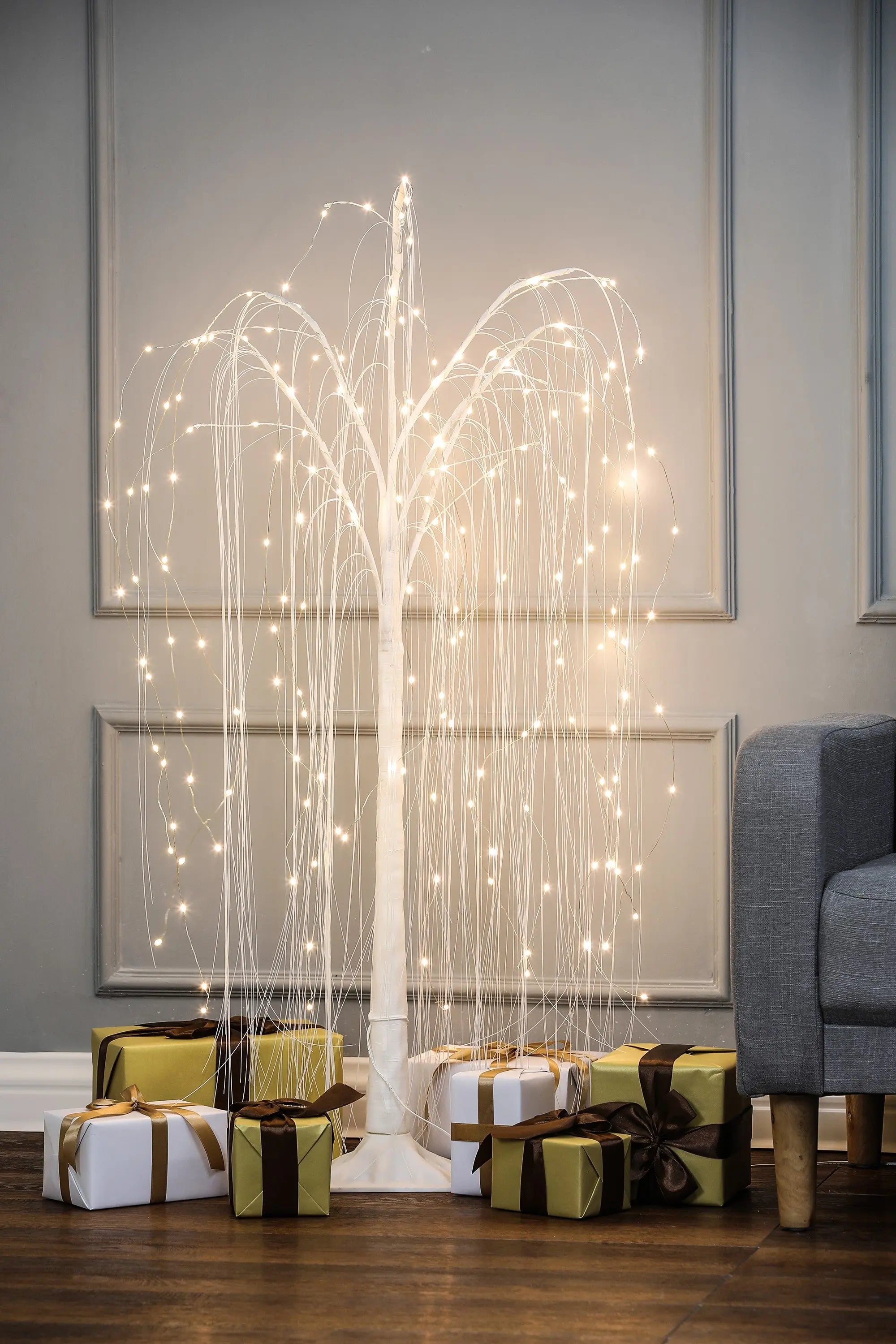 A romantic willow tree with LED lights stands next to a gray sofa, with gold and white wrapped presents arranged underneath on a wooden floor.