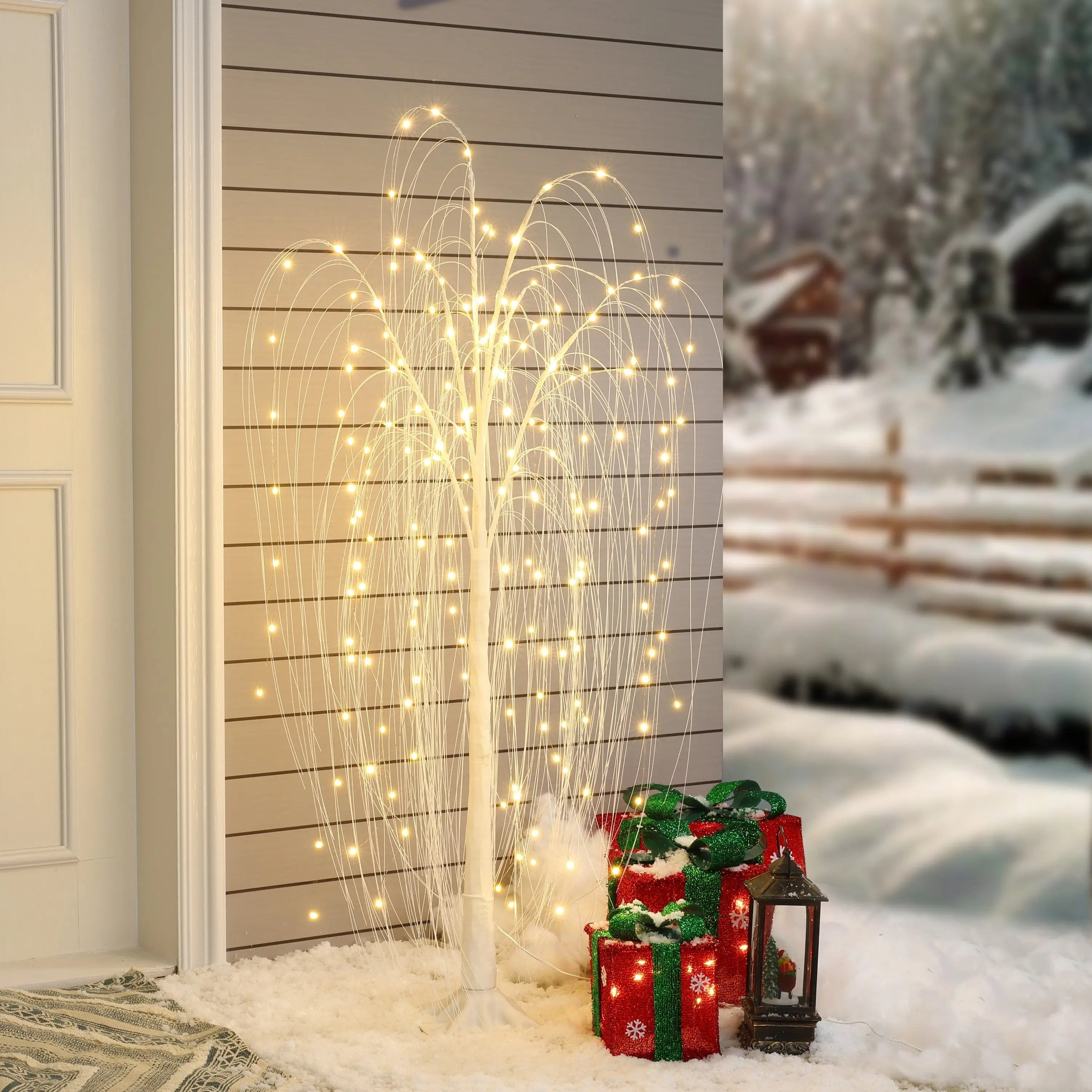 A white pre-lit tree with LED lights stands on snow next to three red holiday gift boxes and a black lantern, set against a snowy outdoor scene for a warm, festive touch.