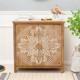 A natural wood finish accent cabinet with white geometric floral scroll work doors, topped with books, decorative birds, and small ornaments, stands next to a plant and vase.