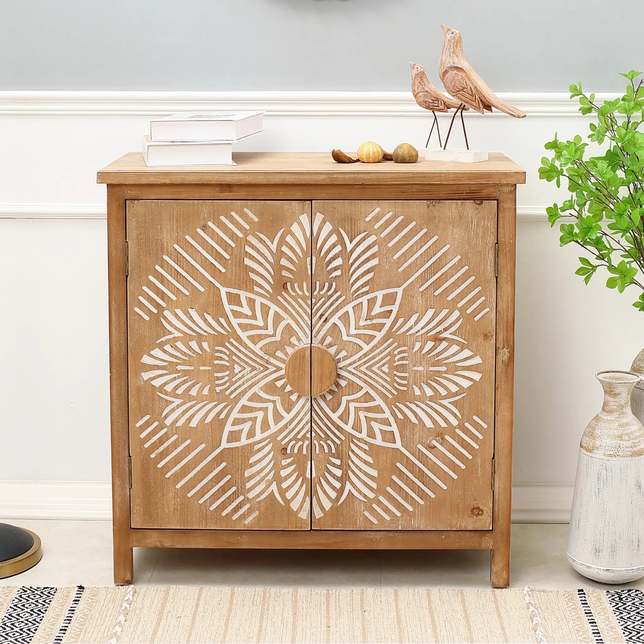 A natural wood finish accent cabinet with white geometric floral scroll work doors, topped with books, decorative birds, and small ornaments, stands next to a plant and vase.