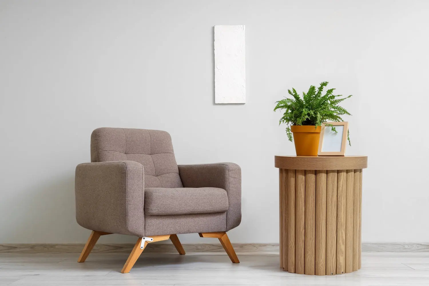 A beige armchair sits beside a round wooden end table with an oak finish, featuring a potted fern and a framed photo against a light gray wall.
