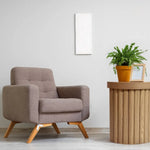A beige armchair sits beside a round wooden end table with an oak finish, featuring a potted fern and a framed photo against a light gray wall.