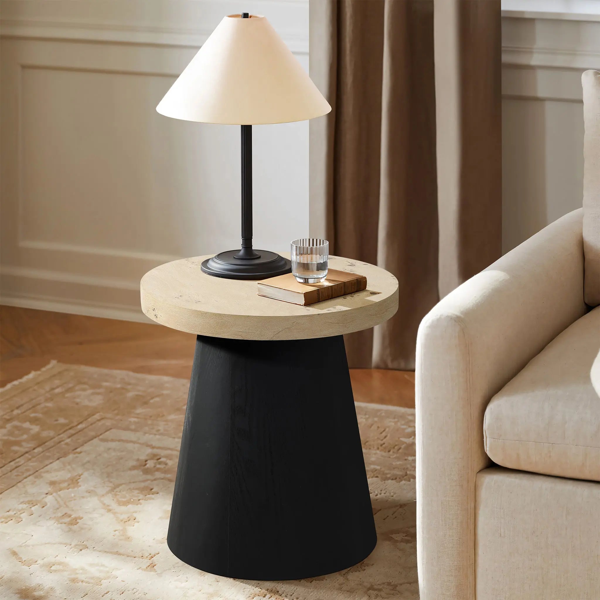 A round side table with a tree burl texture top and black base, featuring a lamp, a stack of books, and a glass cup, stands next to a beige sofa in the living room.