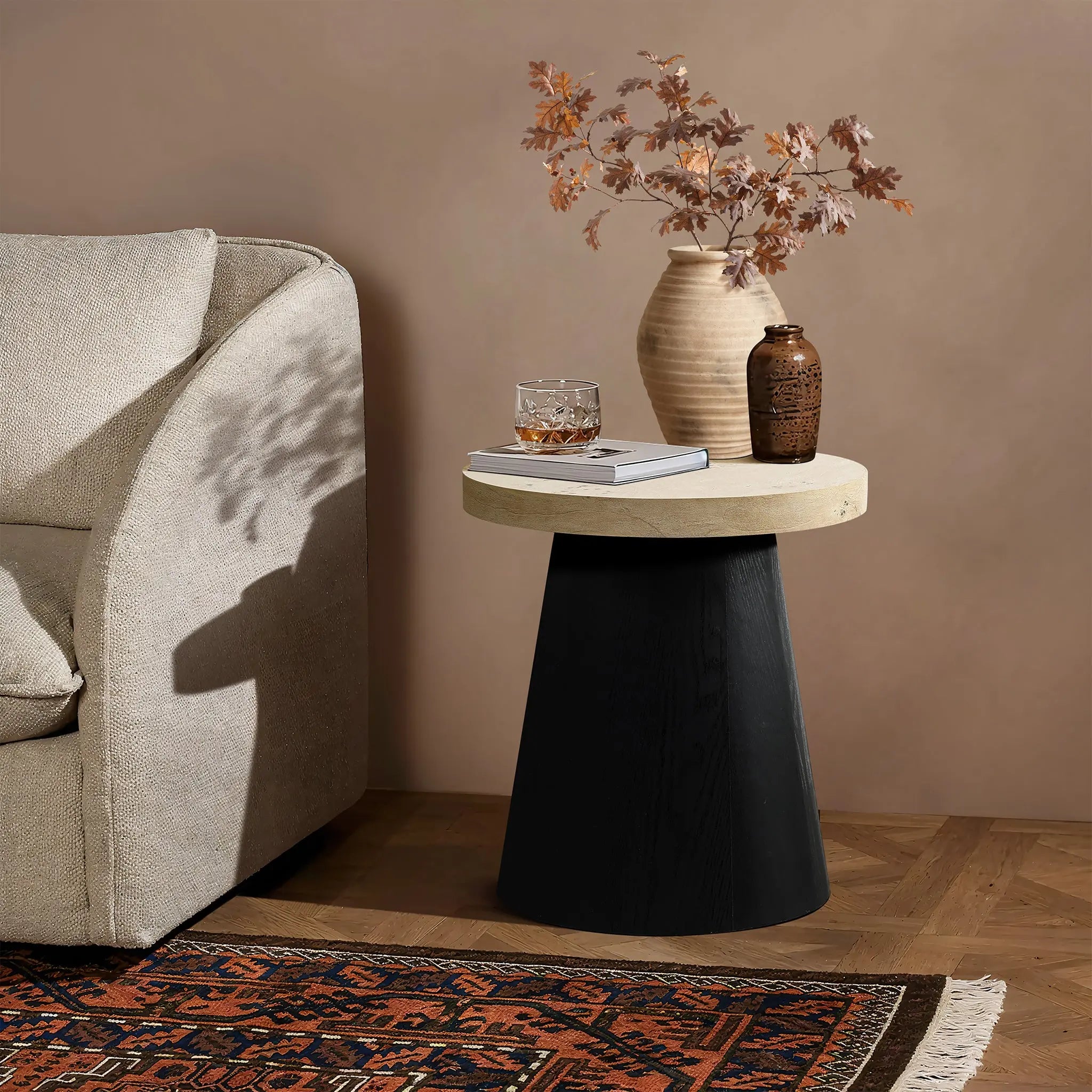 A beige and black side table, crafted from lightweight MDF with a tree burl texture, holds a vase of dried leaves, a small brown jar, a book, and a glass. Next to this elegant setup is a beige sofa resting on a patterned carpeted floor.