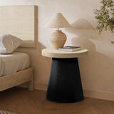 A round side table featuring a tree burl texture and a black base with a cream top sits elegantly beside the bed. It holds a beige lamp and a closed book. The wall and floor showcase neutral tones, while a plant adds life to the background.