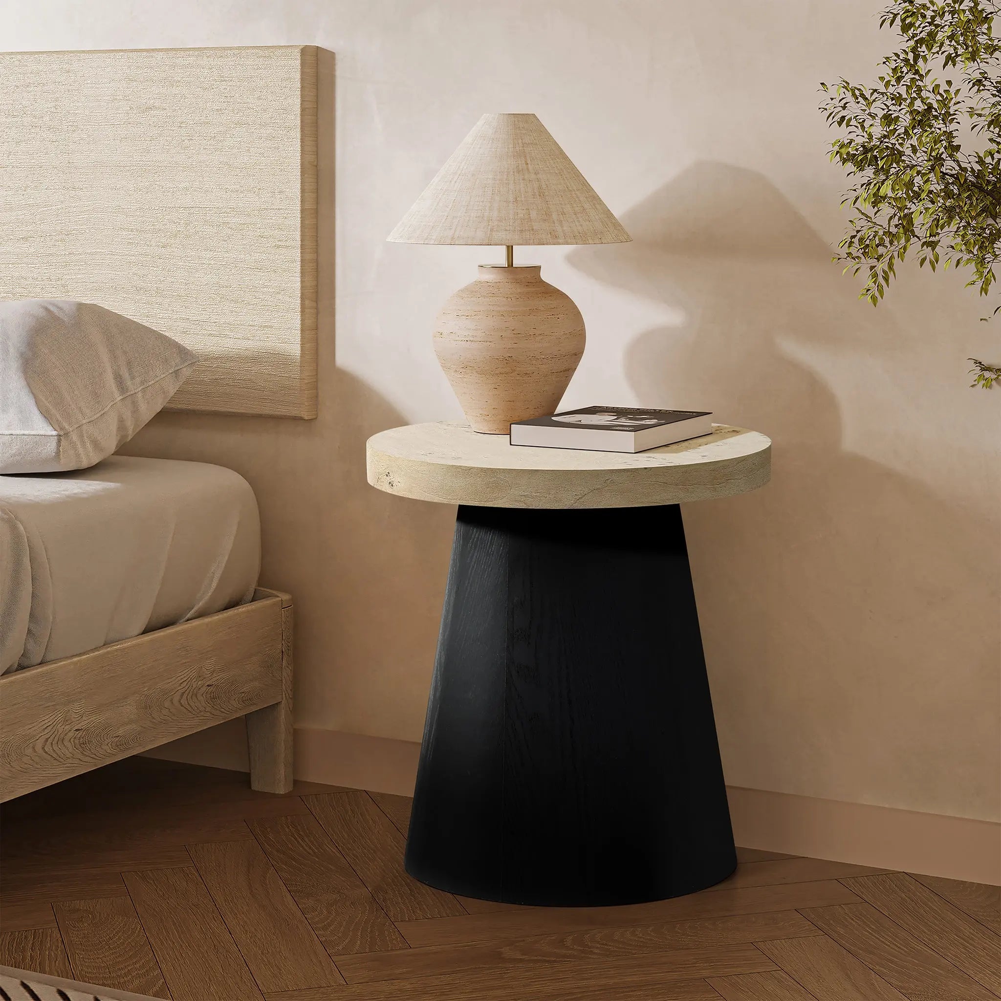 A round side table featuring a tree burl texture and a black base with a cream top sits elegantly beside the bed. It holds a beige lamp and a closed book. The wall and floor showcase neutral tones, while a plant adds life to the background.
