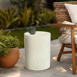 A ceramic mug sits on a round white modern patio table next to a wicker chair with a beige throw blanket; plants are in the background.