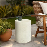 A ceramic mug sits on a round white modern patio table next to a wicker chair with a beige throw blanket; plants are in the background.