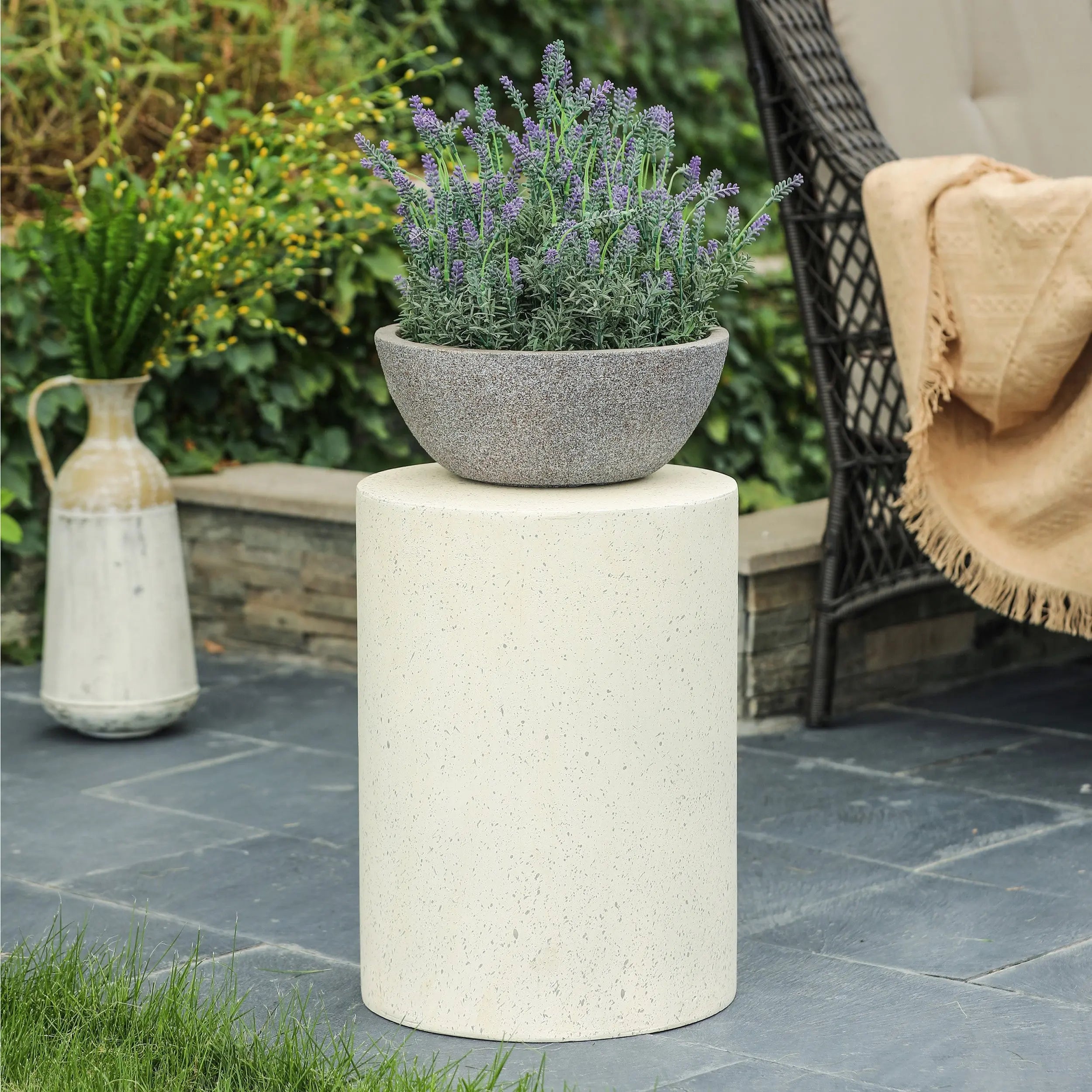 A round white pedestal with a gray stone planter containing purple flowers, set on a gray patio with greenery and modern patio table and outdoor furniture in the background, showcasing contemporary design.