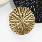 A round, textured, golden disc reminiscent of coastal abstract art sits on a white background, flanked by black and white partially visible discs.
