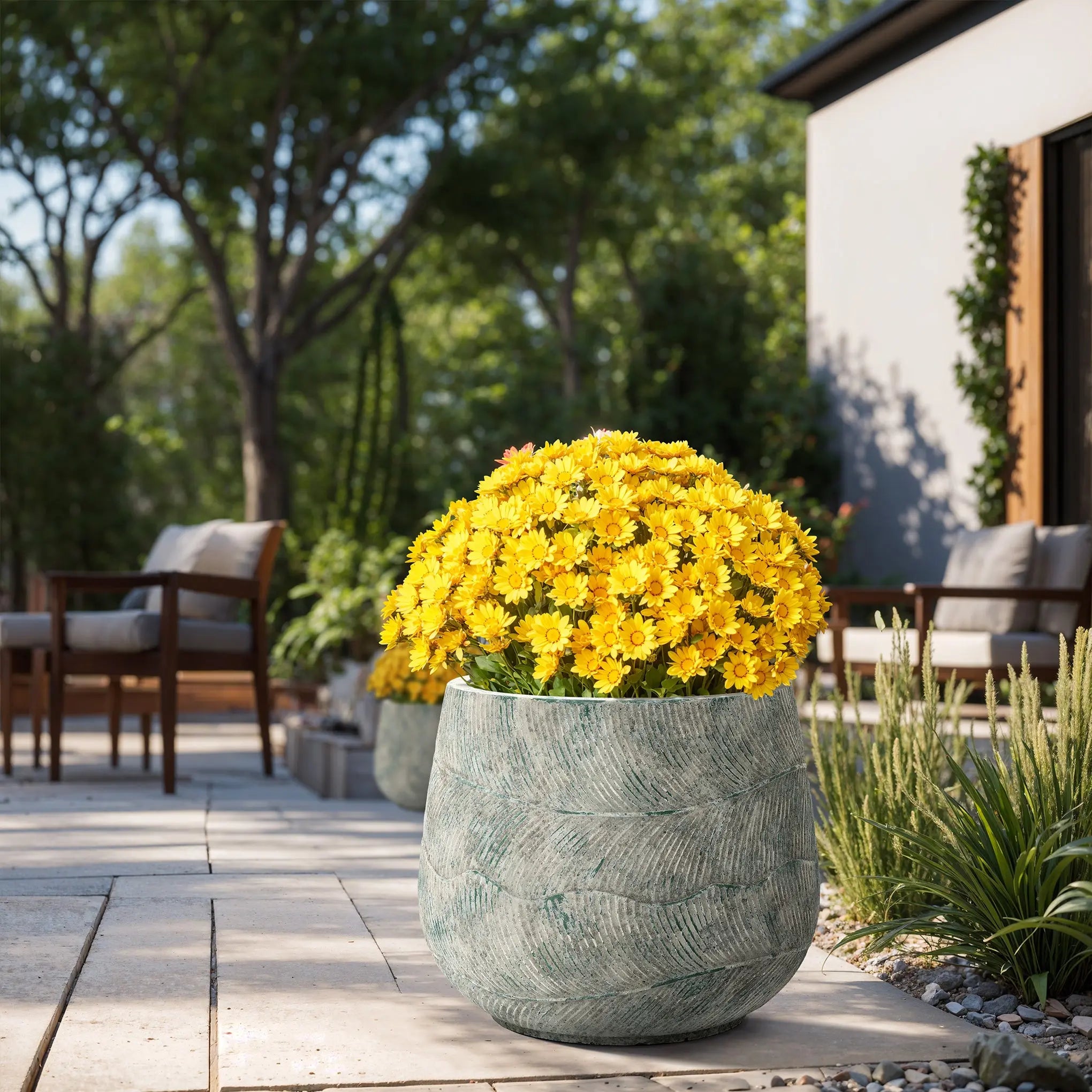 A vibrant outdoor patio displays a LuxenHome Rustic Multi-Color Cement 18.3-Inch Round Planter filled with bright yellow flowers. Wooden chairs with cushions complement the lush greenery and trees under a sunny sky.