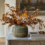 A Rustic brown stoneware bowl vase by LuxenHome with dried eucalyptus branches sits on a gray cabinet. Next to it, books and decorative items are neatly arranged. Above, a framed landscape painting depicting trees and a pastoral scene adds the perfect touch of home decor elegance.