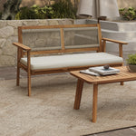 This modern outdoor loveseat set features FSC-certified Acacia wood, showcasing a sleek bench with a white cushion and a matching coffee table. Theyre elegantly arranged on a textured rug atop the stone patio, framed by lush greenery for a serene escape.