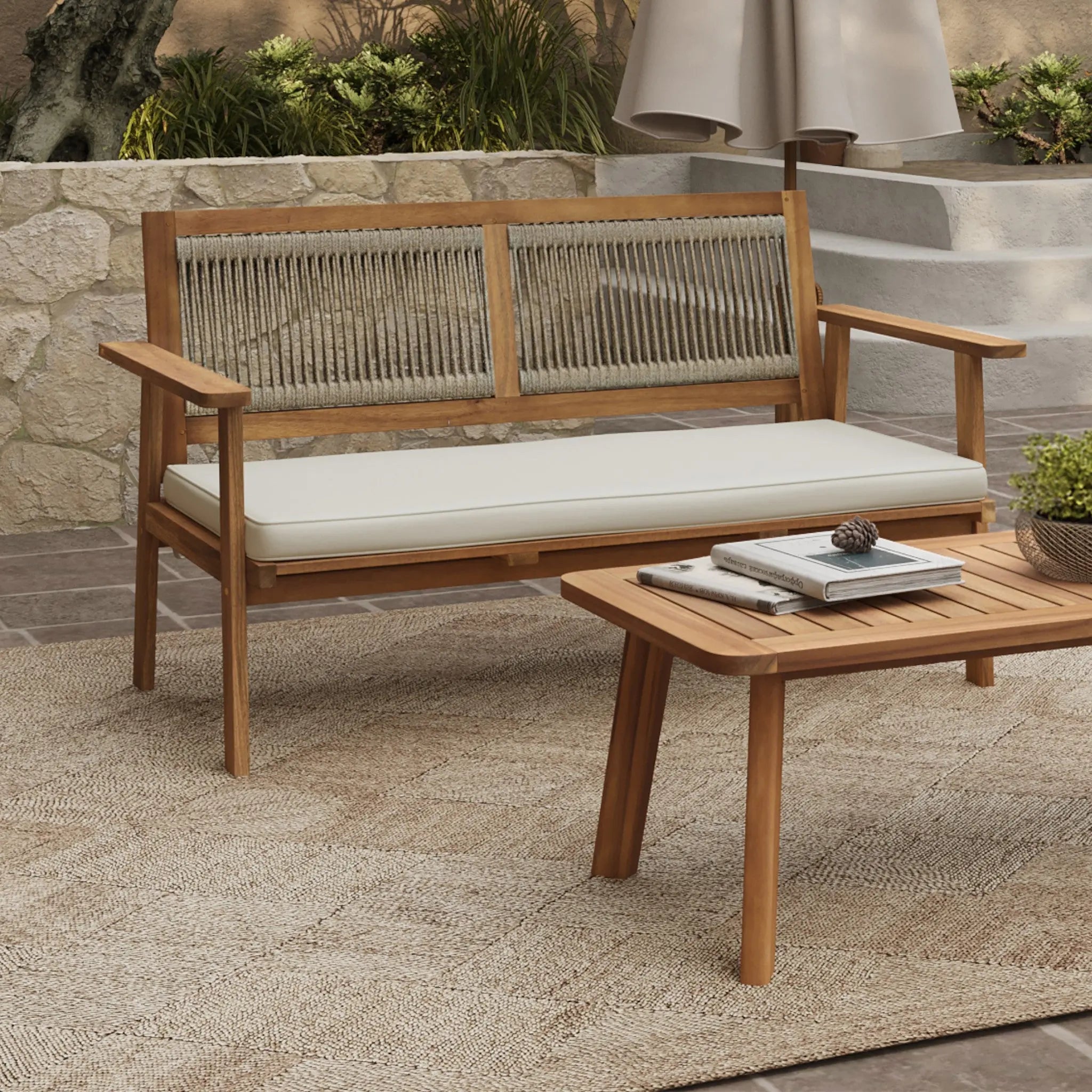 This modern outdoor loveseat set features FSC-certified Acacia wood, showcasing a sleek bench with a white cushion and a matching coffee table. Theyre elegantly arranged on a textured rug atop the stone patio, framed by lush greenery for a serene escape.