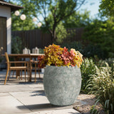 A LuxenHome Rustic Multi-Color Cement 15.4-Inch Round Planter filled with autumn foliage graces a patio, where a wooden table, chairs, and string lights among trees create a serene backyard garden setting.