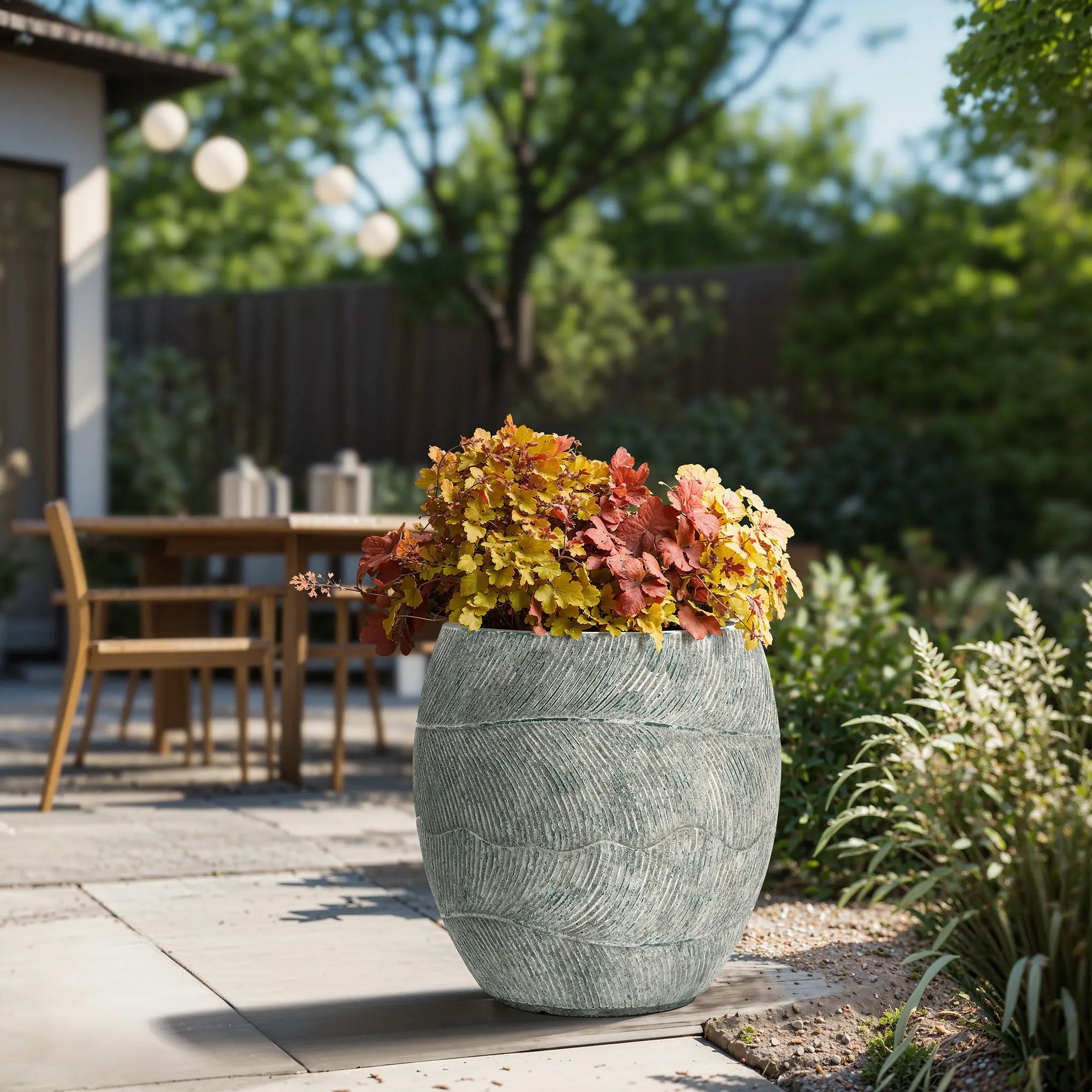A LuxenHome Rustic Multi-Color Cement 15.4-Inch Round Planter filled with autumn foliage graces a patio, where a wooden table, chairs, and string lights among trees create a serene backyard garden setting.