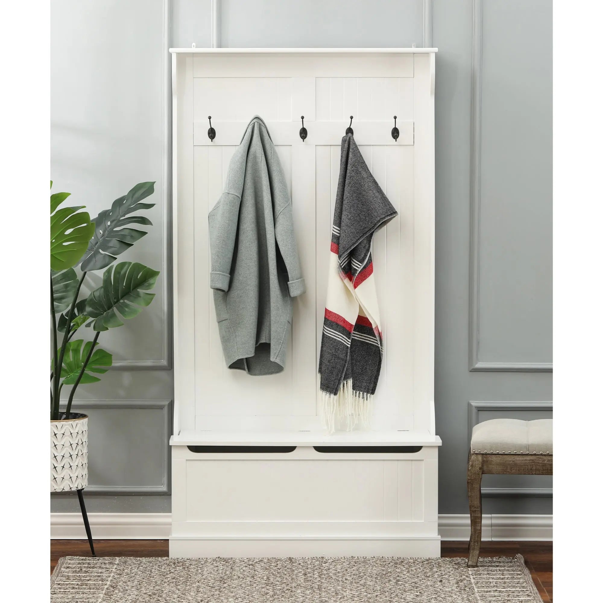 A white hall tree with black hooks serves as a coat rack, holding a gray coat and a striped scarf. A potted plant, storage bench, and gray rug are nearby.