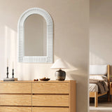 A sunlit bedroom features a wooden dresser topped with stylish home decor, including candles and small trinkets. An ornate wall mirror complements the table lamp, while the bed with white bedding peeks out from the background, adding to the rooms elegant charm.