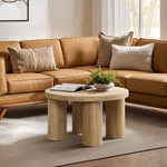 A round, oak-finish coffee table with a plant and an open book graces the space in front of a tan sectional sofa adorned with cushions, all bathed in natural light.