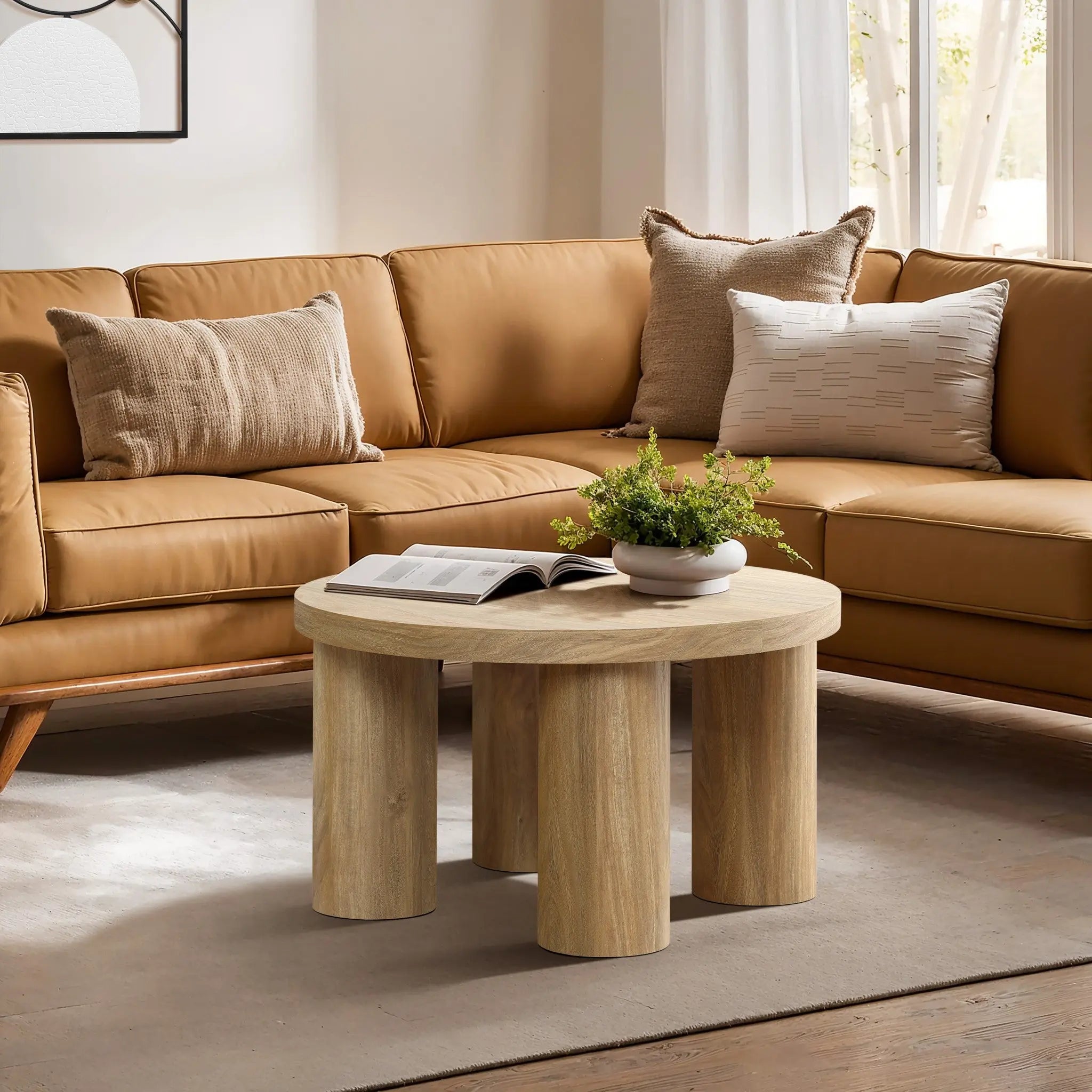 A round, oak-finish coffee table with a plant and an open book graces the space in front of a tan sectional sofa adorned with cushions, all bathed in natural light.