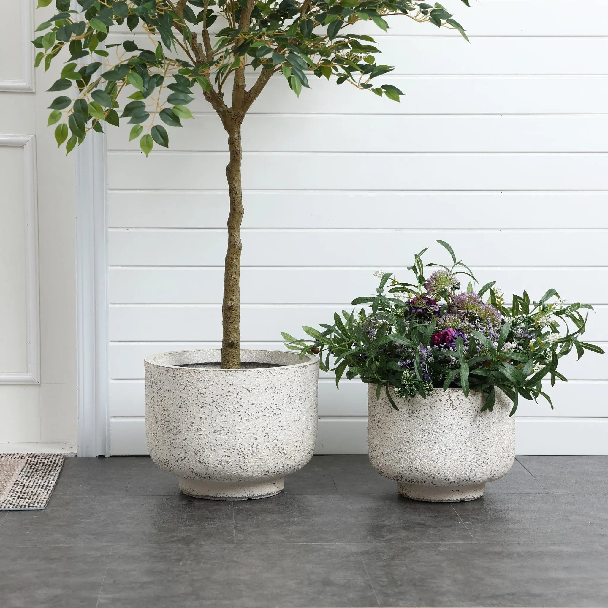 Two large white ceramic pots grace the gray tile floor, one with a small tree and the other bursting with green plants and purple flowers, against a white paneled wall. Their weather-resistant design speaks to retro minimalist decor, making them stylish yet practical.