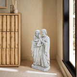 A marble-like polyresin Holy Family statue stands in a corner, flanked by a wooden cabinet on the left and a large window on the right, resting gracefully on a light beige carpeted floor.