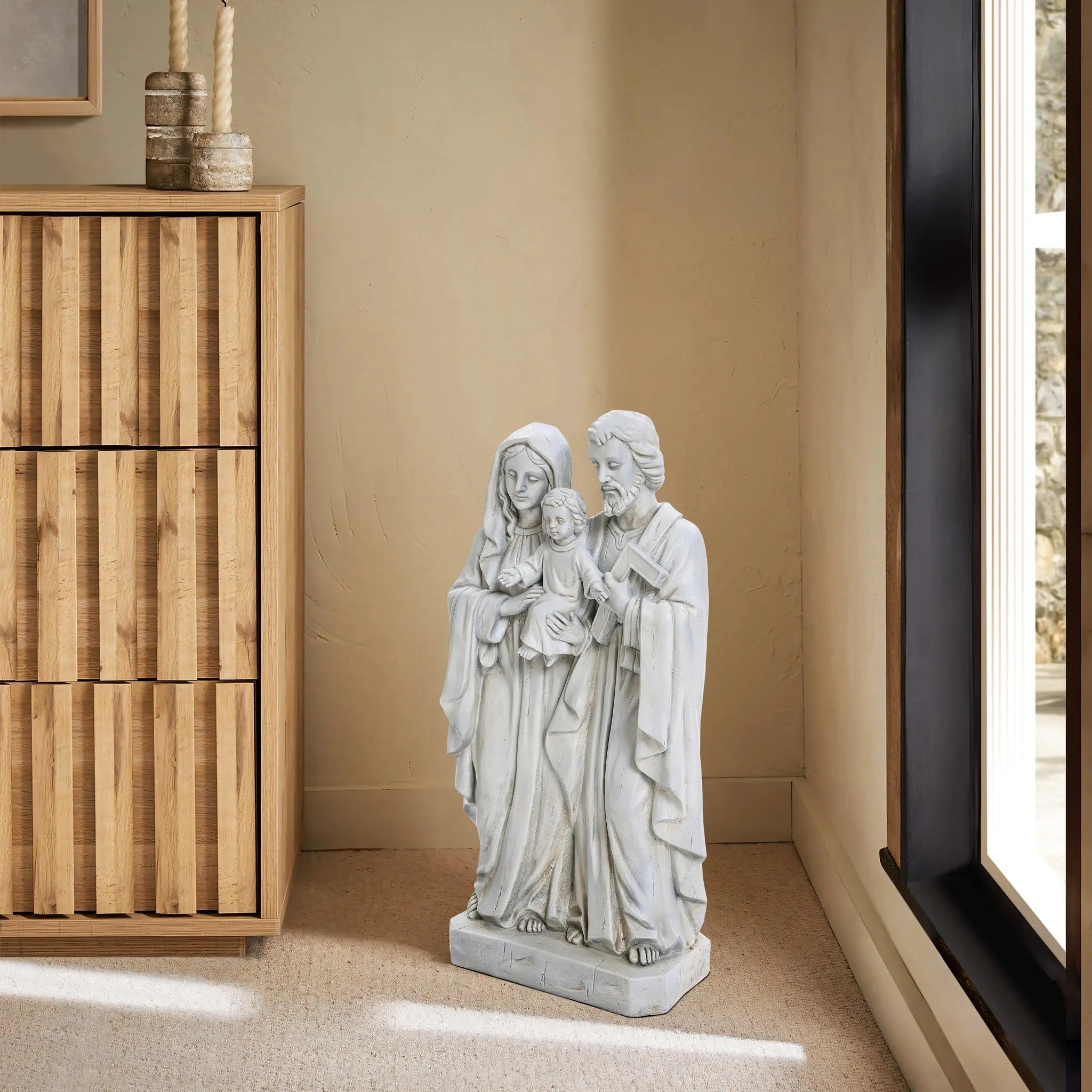 A marble-like polyresin Holy Family statue stands in a corner, flanked by a wooden cabinet on the left and a large window on the right, resting gracefully on a light beige carpeted floor.
