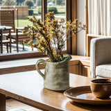A ceramic pitcher vase adorned with yellow flowers sits on a wooden table near a window, its artisanal craftsmanship evident. Nearby, a brown bowl and plate complete the setting as sunlight illuminates the scene.