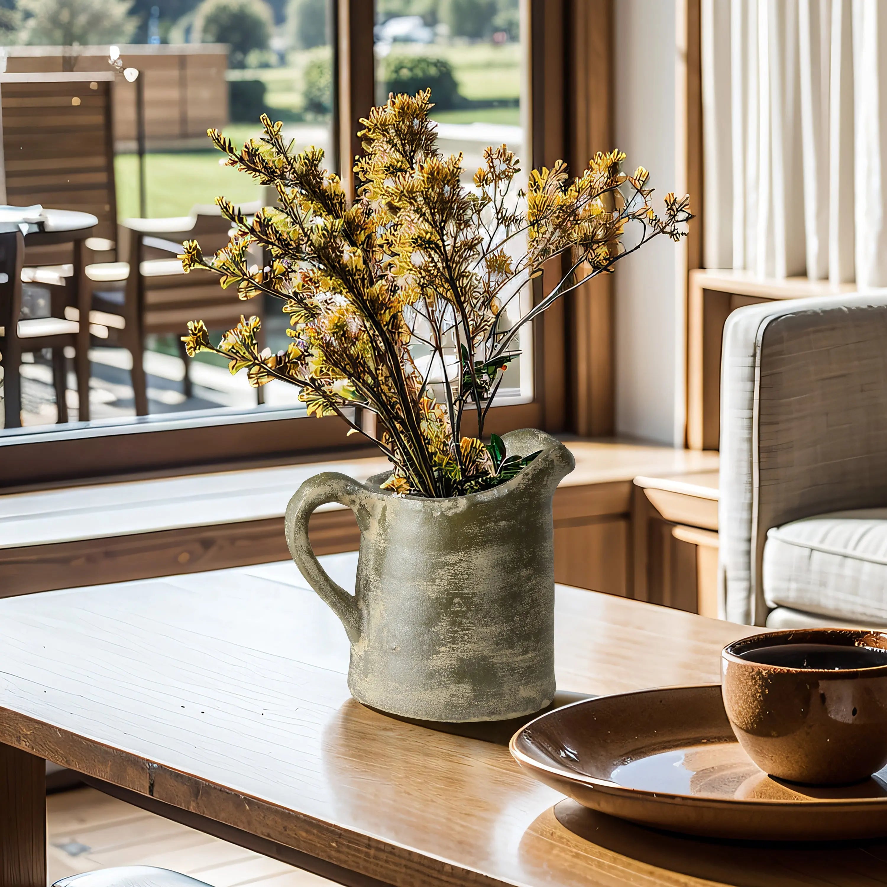 A ceramic pitcher vase adorned with yellow flowers sits on a wooden table near a window, its artisanal craftsmanship evident. Nearby, a brown bowl and plate complete the setting as sunlight illuminates the scene.