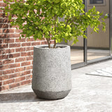 Gray stone planter with a leafy green plant on a concrete surface, showcasing a lightweight durable MgO construction. It features a gray and white speckled texture, complementing its modern design against the red brick wall and glass doors.