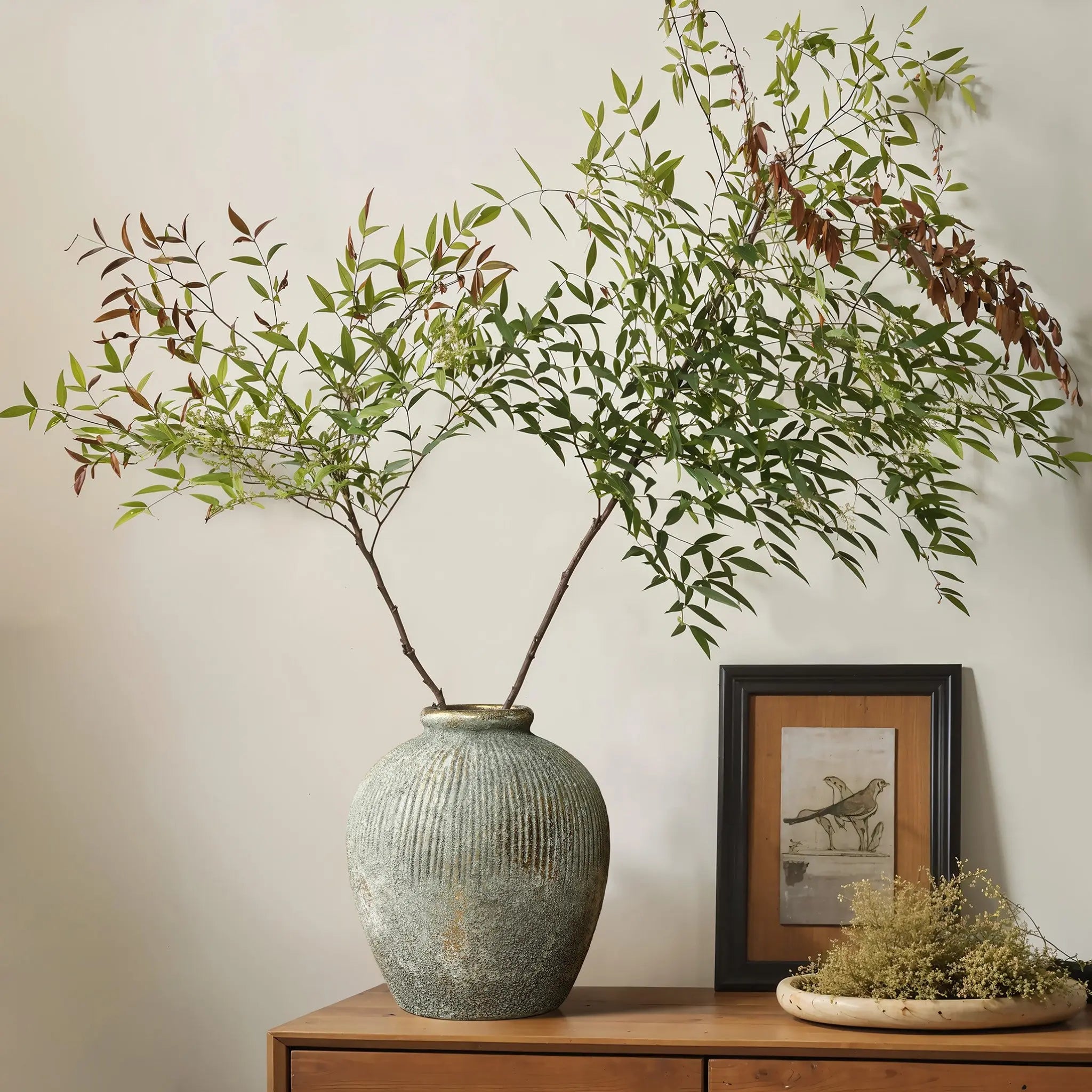 A LuxenHome rustic gold and brown terracotta bud vase (8.7 h) adorns the wooden sideboard alongside a small framed bird illustration and a shallow dish with dried flowers, elevating the home decor against an antique distressed neutral-colored wall.