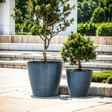 Two decorative planters with a UV-protected finish are placed on a paved area outdoors, housing lush potted plants. The large gray planters seamlessly complement the greenery and benches in the background, perfect for both indoor and outdoor use.